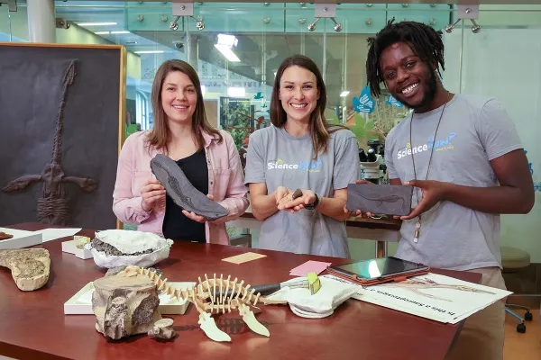 Two light-skinned women and one dark-skinned man holding fossils while standing behind a table with fossils on it, including what looks like a model of a skeleton of a long-necked creature with flippers. A large fossil of a similar creature embedded in stone stands upright to the left of the people.