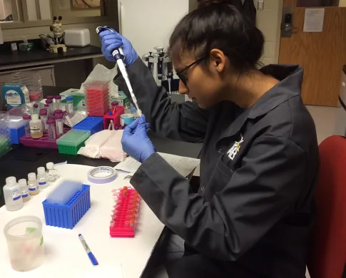 A teenage girl wearing a black jacket and blue gloves sitting at a lab table. She is holding a pipette in her right hand and inserting it into a test tube in her left hand.