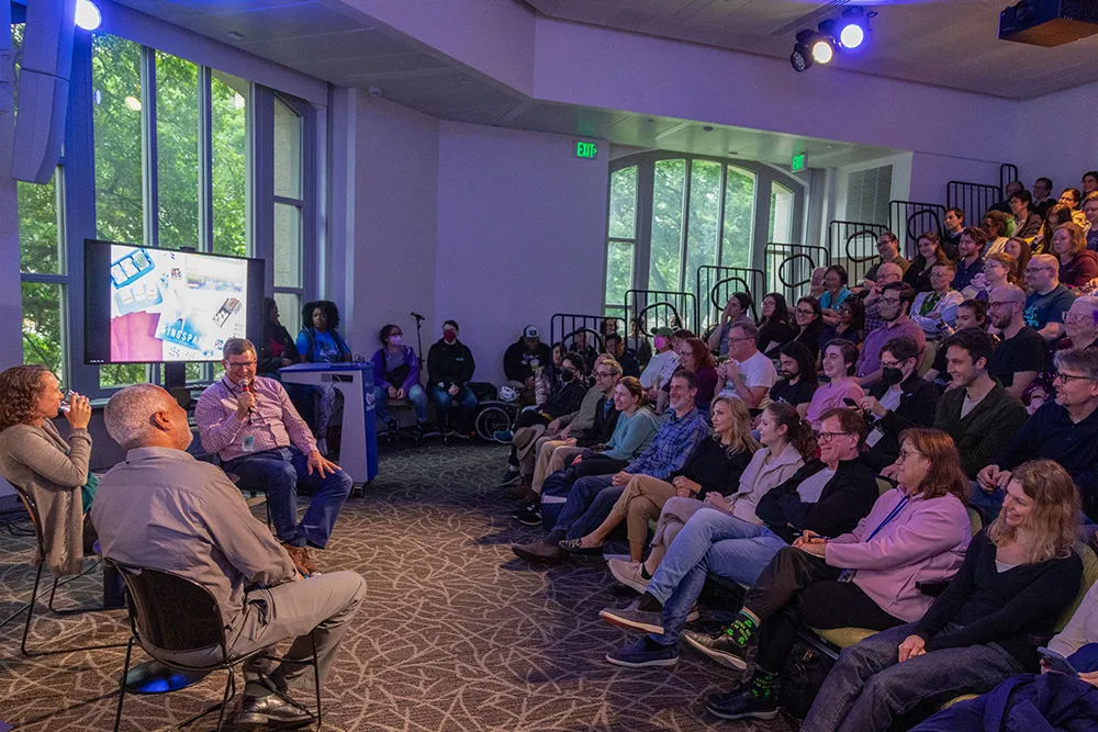 A scene inside a small theater. At left, three people sit on chairs on the floor in front of about 100 people sitting in auditorium-style seating on the right. One of the seated people has a microphone and there is a video monitor behind him.