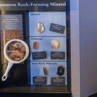Exhibit case titled Common Rock-Forming Minerals. Three sections on the right contain specimens of quartz, feldspar, and iron-rich minerals. On the left is an oversized magnifying glass showing a zoomed-in image of a rock with its mineral components labeled.