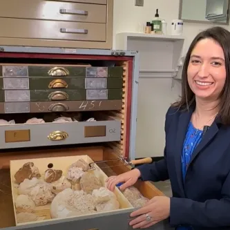 A woman with shoulder-length brown hair, wearing a navy blue blazer over a royal blue shirt, stands next to an open drawer of coral specimens. The drawer is one of many in a cabinet of drawers.